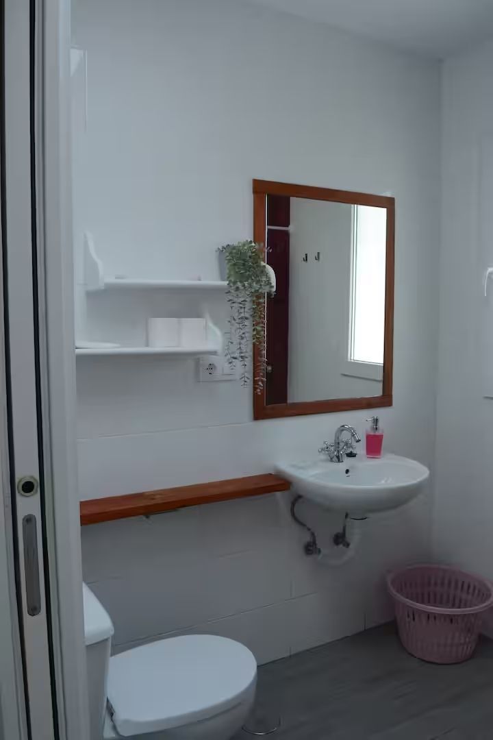 Baño con paredes blancas, inodoro, lavabo, espejo, estantería con decoración y una cesta rosa.