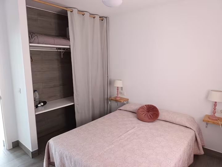 Dormitorio con ropa de cama rosa, una almohada redonda y un armario con cortinas.