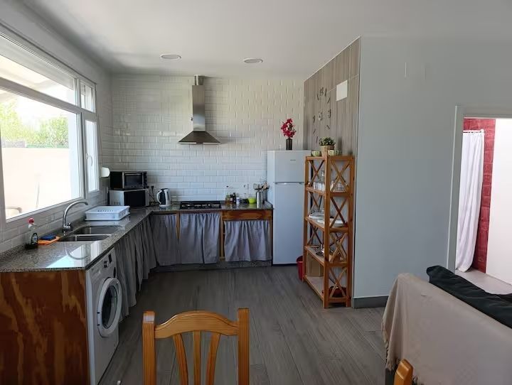 Cocina con lavadora, fregadero y nevera, en tonos grises y blancos. Estantes de madera y mesa de comedor.