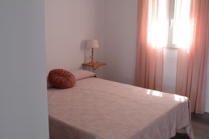 Dormitorio con cama rosa, cortinas y almohada redonda. Mesa pequeña con lámpara cerca de la ventana.