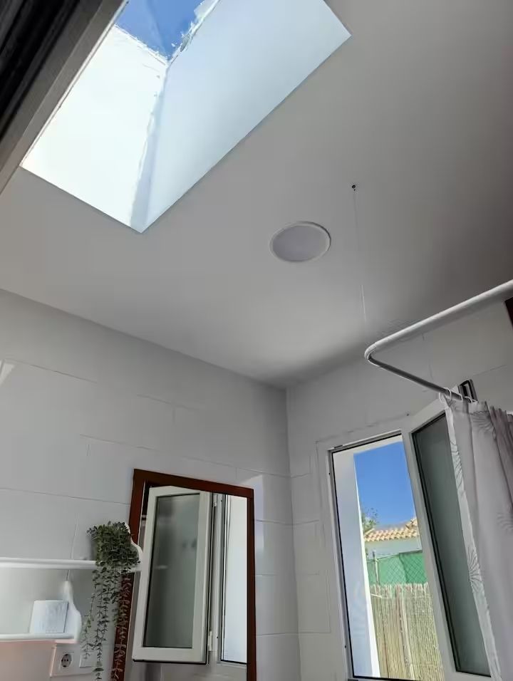 Interior de baño con claraboya, ventana y espejo. Paredes blancas y luz natural.