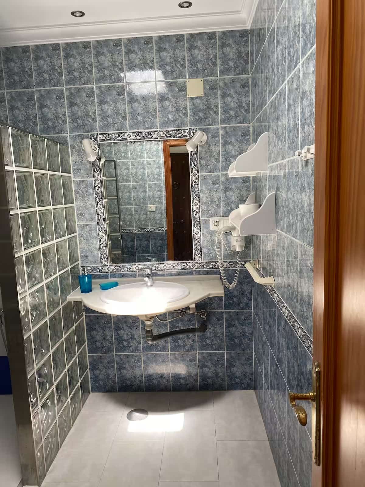 Baño con paredes de azulejos azules, lavabo con espejo y ducha de bloques de vidrio.