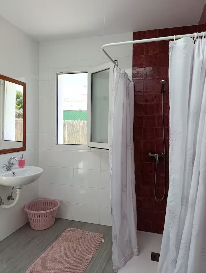 Baño con paredes blancas, ventana, pared de ducha roja y accesorios rosas.