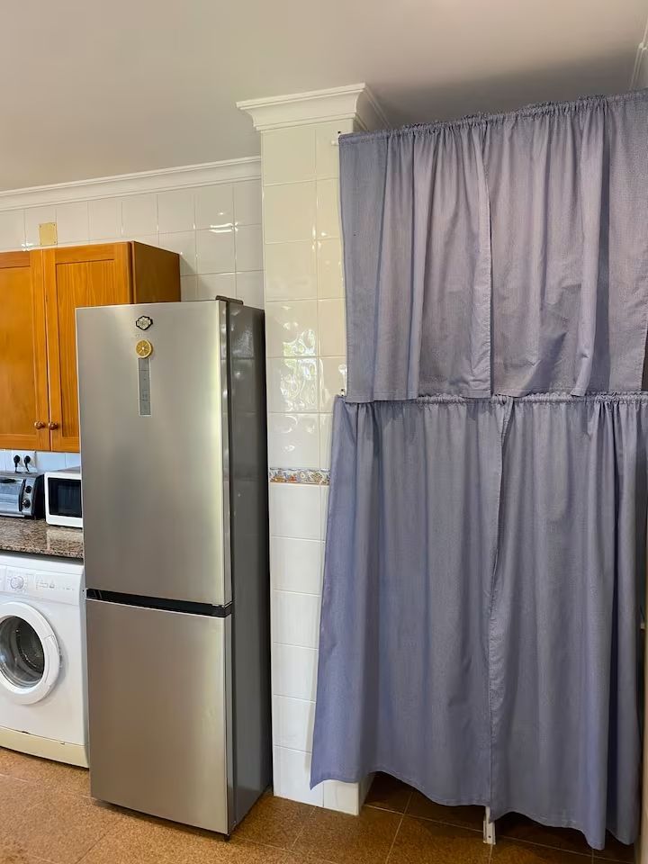 Una cocina con refrigerador, lavadora, gabinetes de madera y una cortina azul.