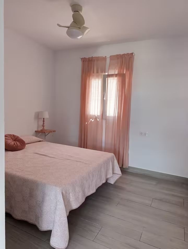 Dormitorio con cama, ventana con cortinas rosas y ventilador de techo.