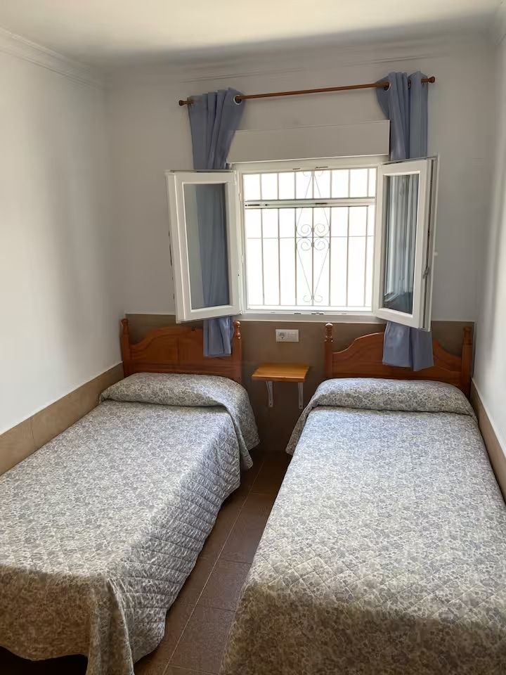 Dos camas individuales en una habitación blanca con una ventana abierta, cortinas azules y una pequeña mesita de noche.