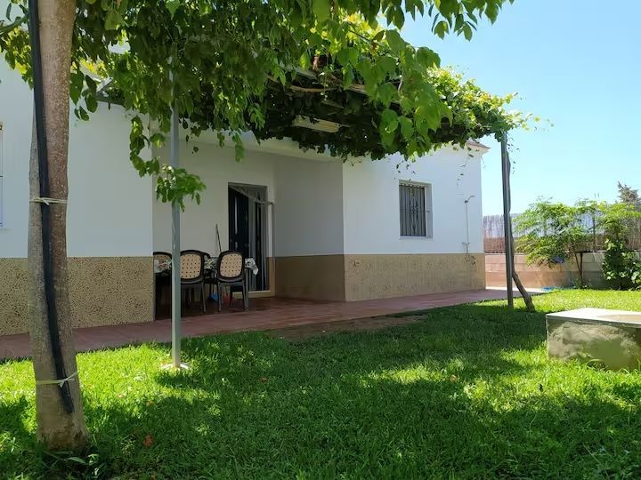 Una casa blanca con enredaderas verdes en un enrejado, tres sillas en un porche y un patio con césped.