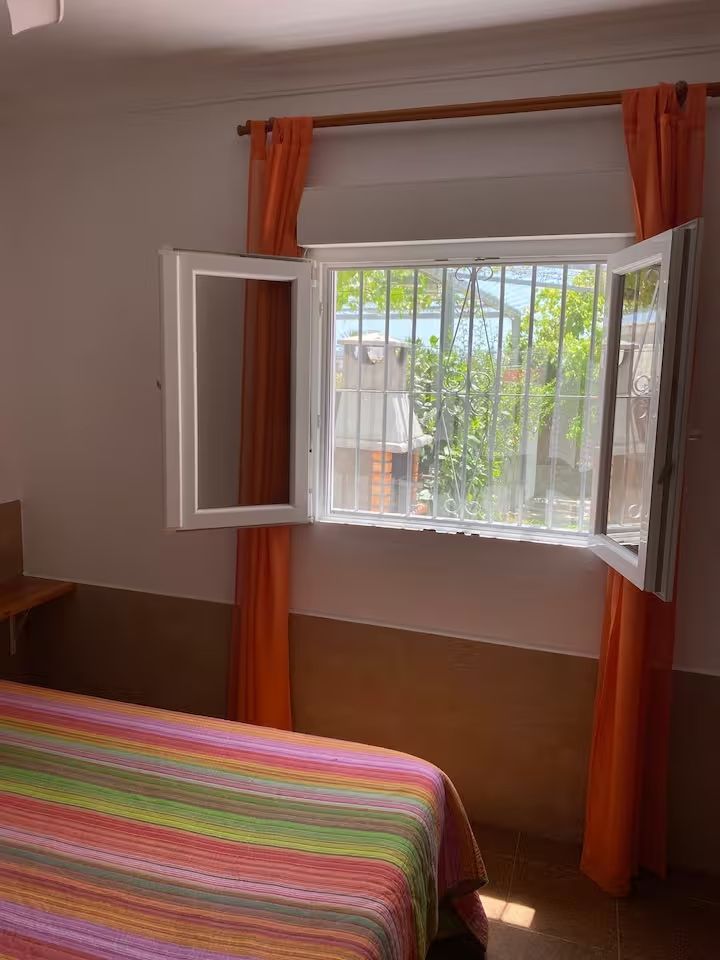 Ventana abierta con cortinas naranjas, cama con colcha de rayas y vista a la vegetación.