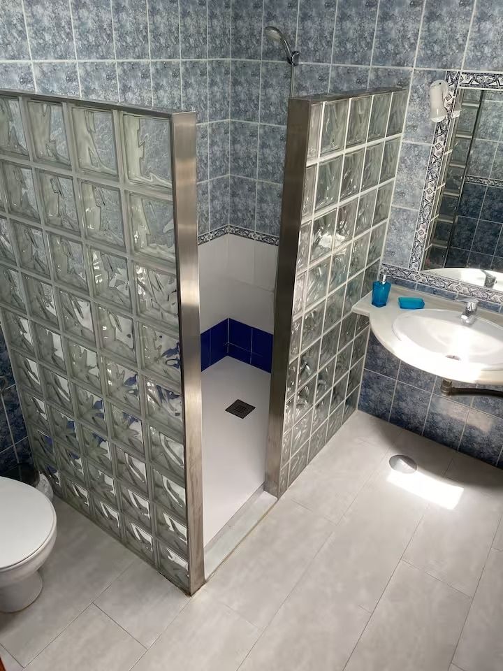 Baño con mampara de ducha de bloques de vidrio, paredes de azulejos azules, lavabo blanco e inodoro.