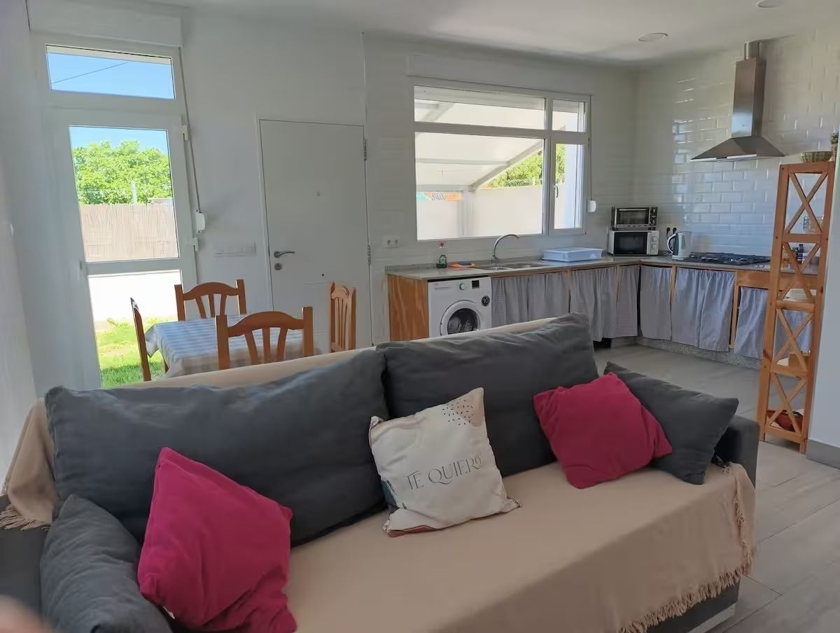 Sala de estar y cocina con sofá gris y cojines rosas, paredes blancas brillantes.