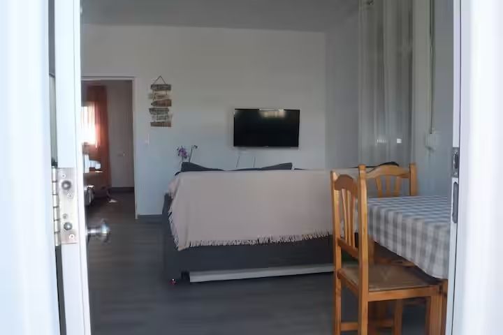 Puerta abierta a un pequeño estudio: cama, televisor, mesa y sillas. Colores neutros con luz natural.