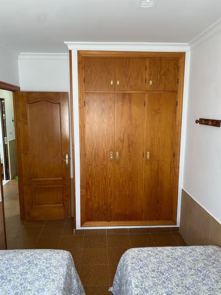 Armario de madera y puerta cerrada en dormitorio con dos camas individuales, suelo de baldosas marrones, molduras blancas.