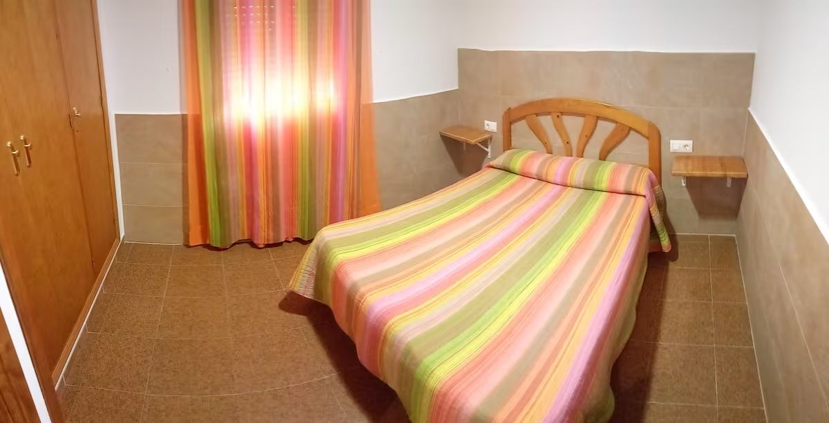 Dormitorio con cama, mesitas de noche, armario y cortinas de rayas en tonos rosa, verde y naranja.