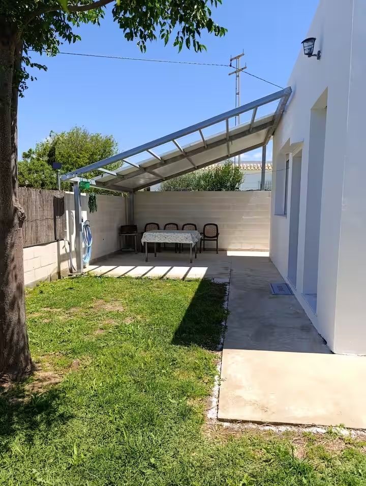 Patio trasero con toldo metálico, mesa, sillas y césped verde. Un día soleado y luminoso.