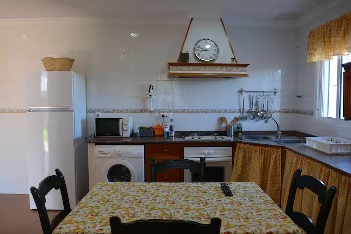 Cocina con mesa, electrodomésticos y reloj. Incluye refrigerador blanco, encimera oscura y cortinas amarillas.