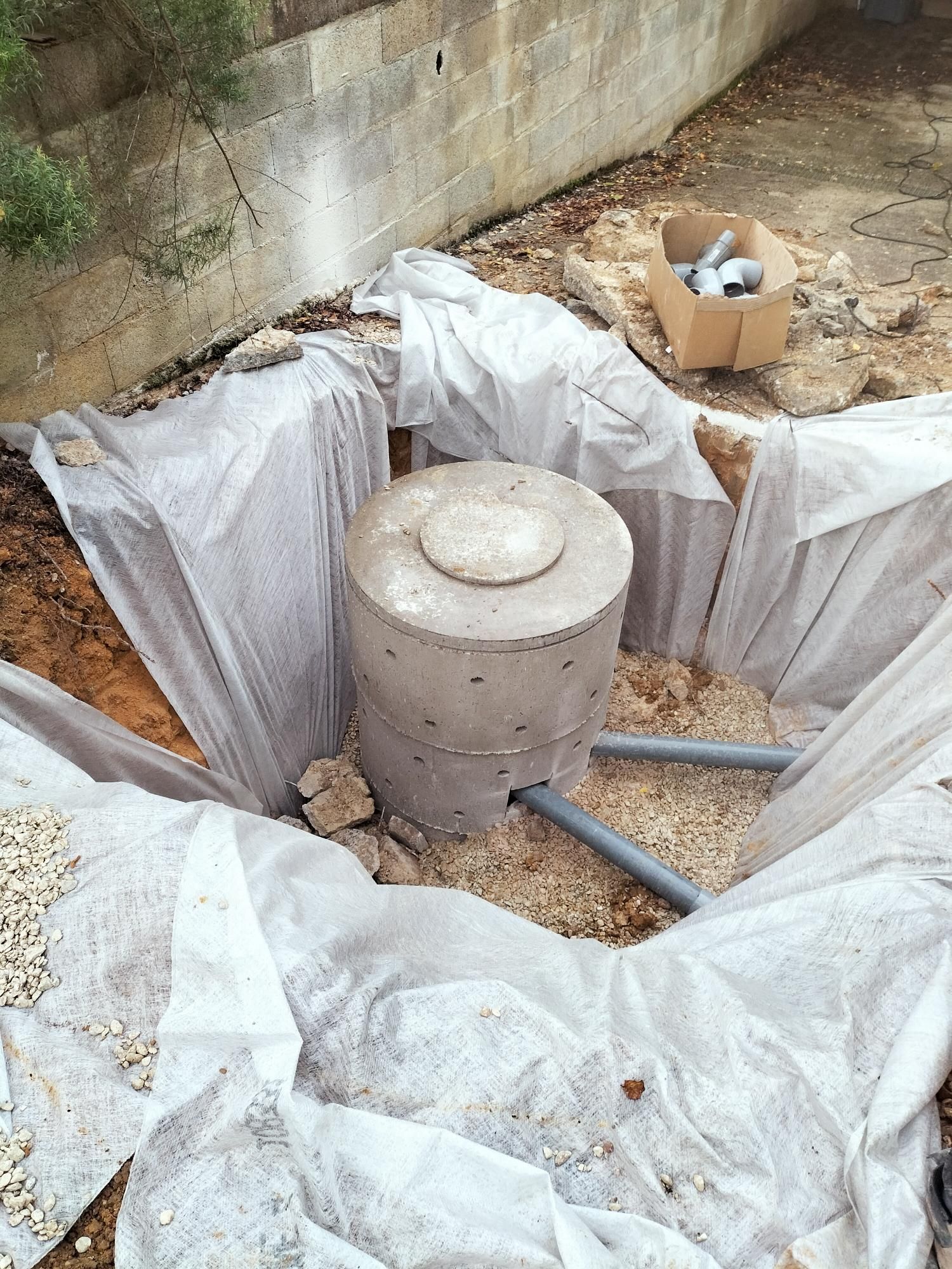 Fosse à ciel ouvert tapissée d’une bâche blanche, contenant un cylindre en béton et une boîte en carton, sur une pente de terre
