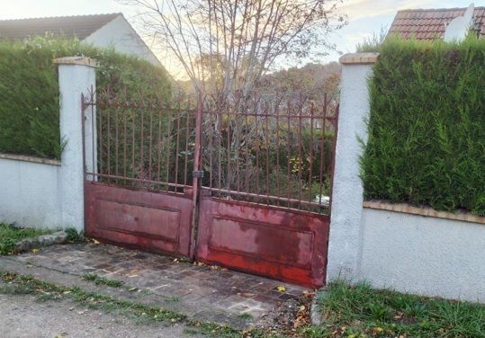 Portail métallique rouge entre des murs blancs et des haies vertes, à l’entrée d’une allée