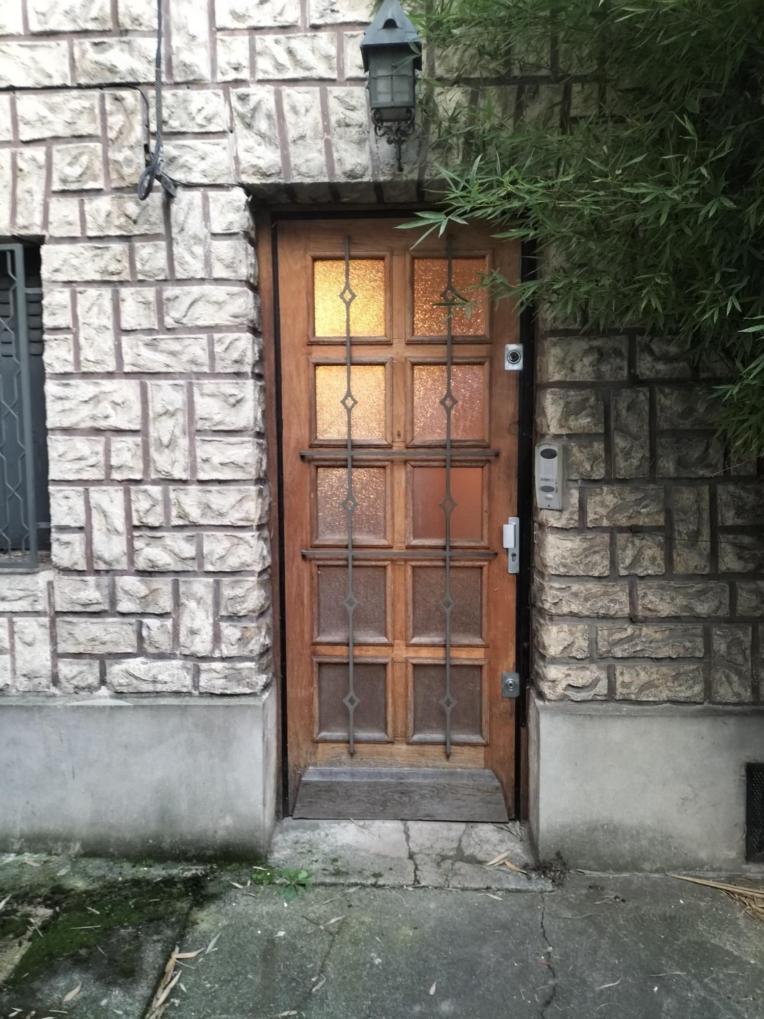 Porte d’entrée en bois dans un mur de pierre, avec une lanterne au-dessus et des marches en béton en dessous
