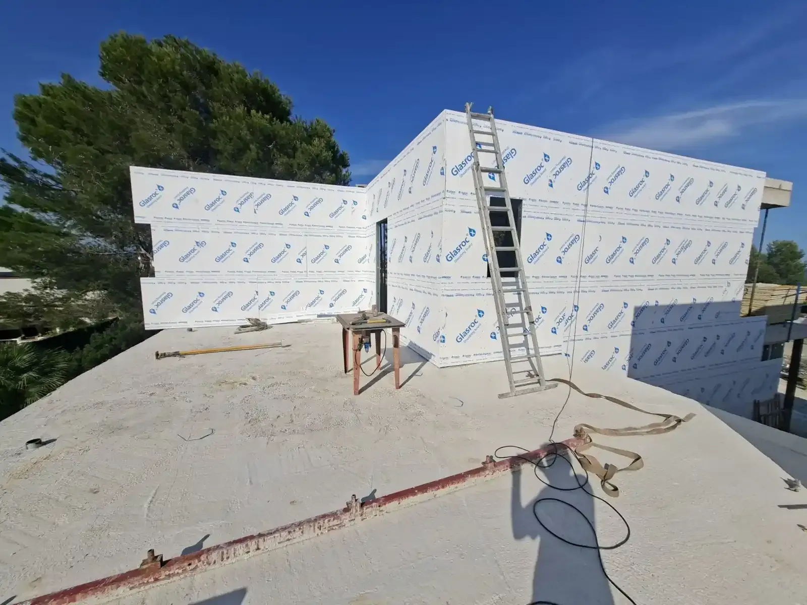 Casa parcialmente en construcción con escalera y manguera en el tejado bajo un cielo azul.