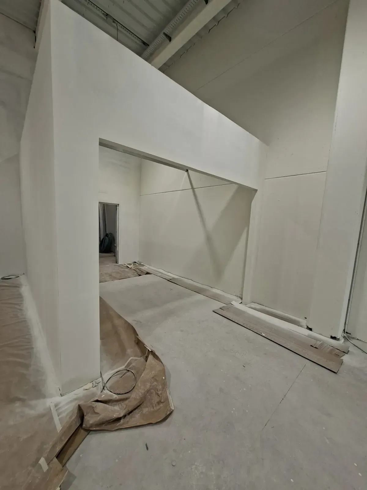 Interior blanco sin terminar, con paredes en ángulo, vigas a la vista y materiales de construcción en el suelo.