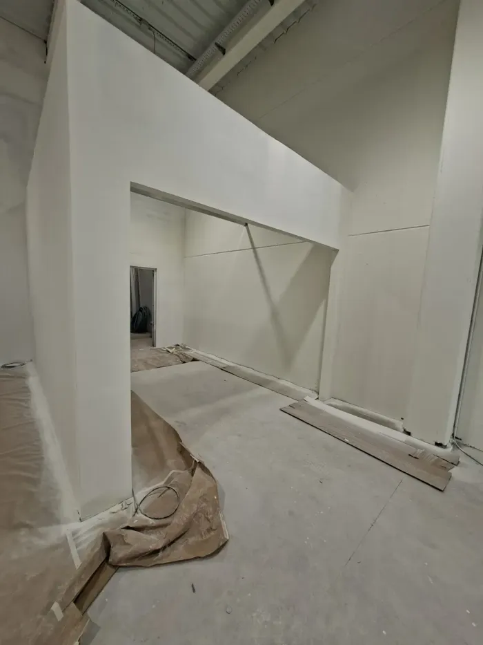 Interior blanco sin terminar, con paredes en ángulo, vigas a la vista y materiales de construcción en el suelo.