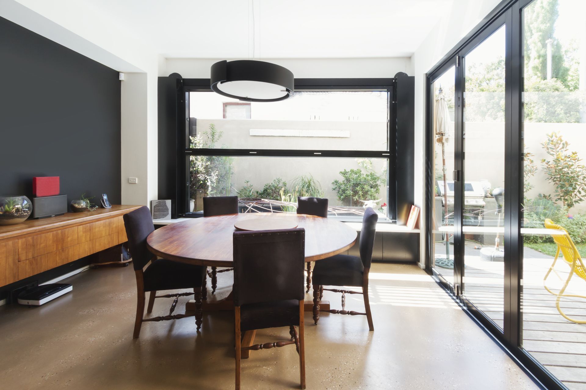 Salle à manger moderne avec table ronde en bois, chaises foncées, grandes fenêtres et portes vitrées donnant sur une terrasse