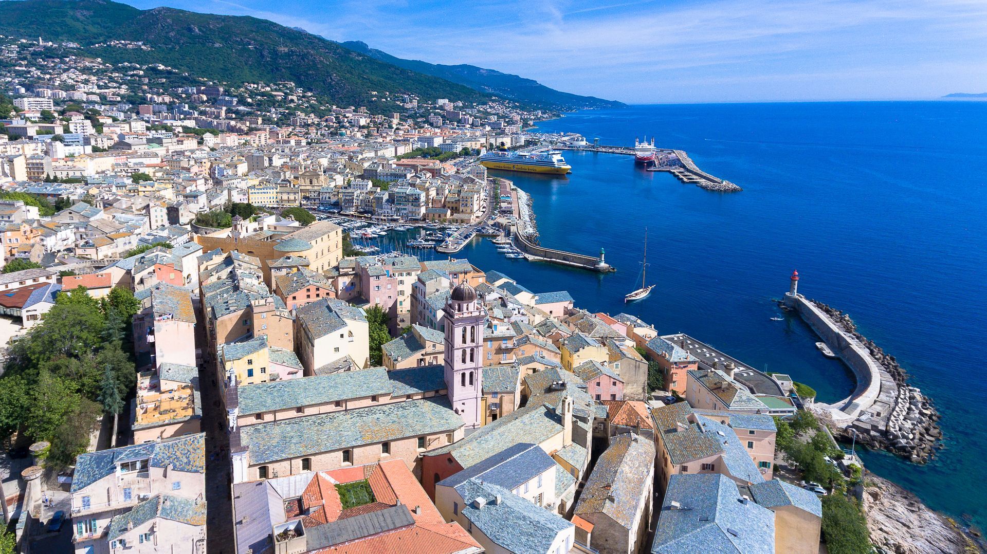Vue aérienne d'une ville côtière avec des bâtiments colorés, un port et une mer bleue sous un ciel ensoleillé