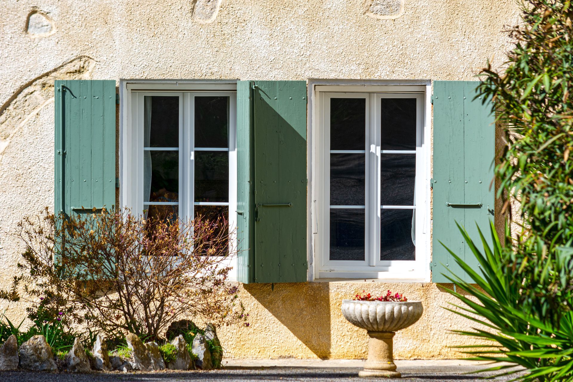 Une façade de maison en stuc avec des volets bleu sarcelle, deux fenêtres, des fleurs en pot et un jardin ensoleillé.