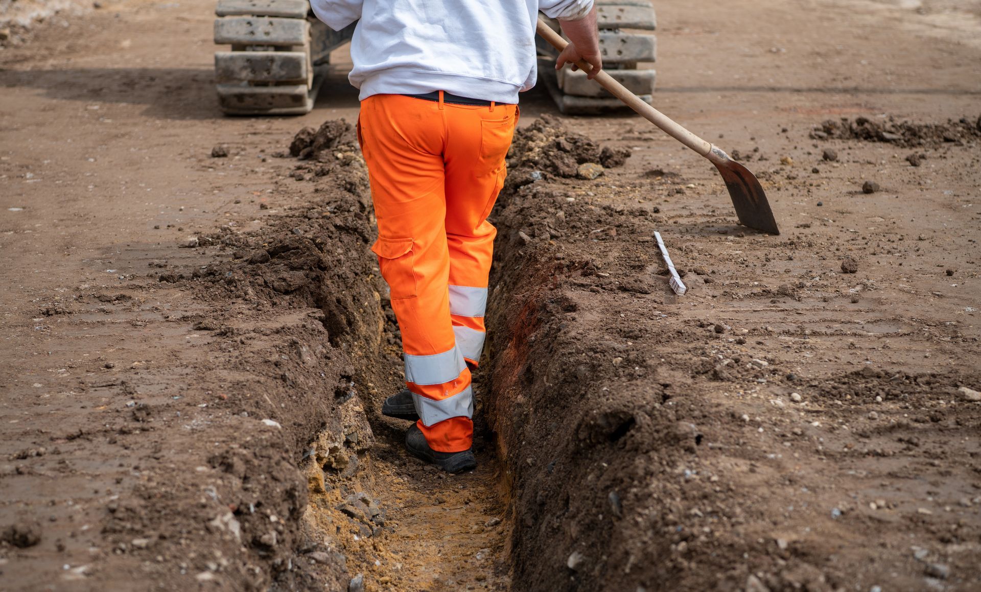 Un ouvrier en pantalon de sécurité orange creuse une tranchée à la pelle sur un chantier de construction.