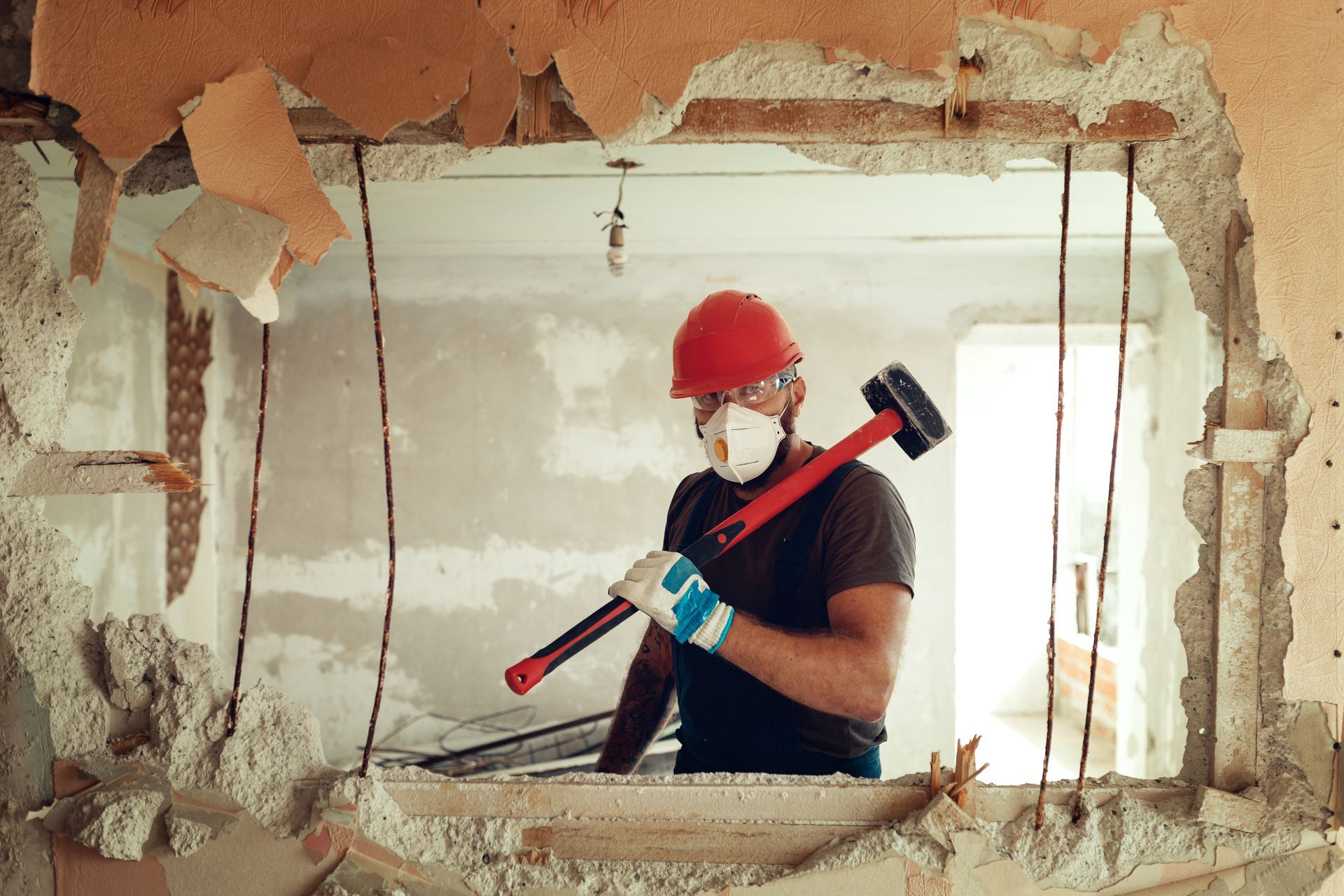 Un ouvrier portant un casque rouge et un masque utilise une masse pour démolir un mur lors de travaux de rénovation.