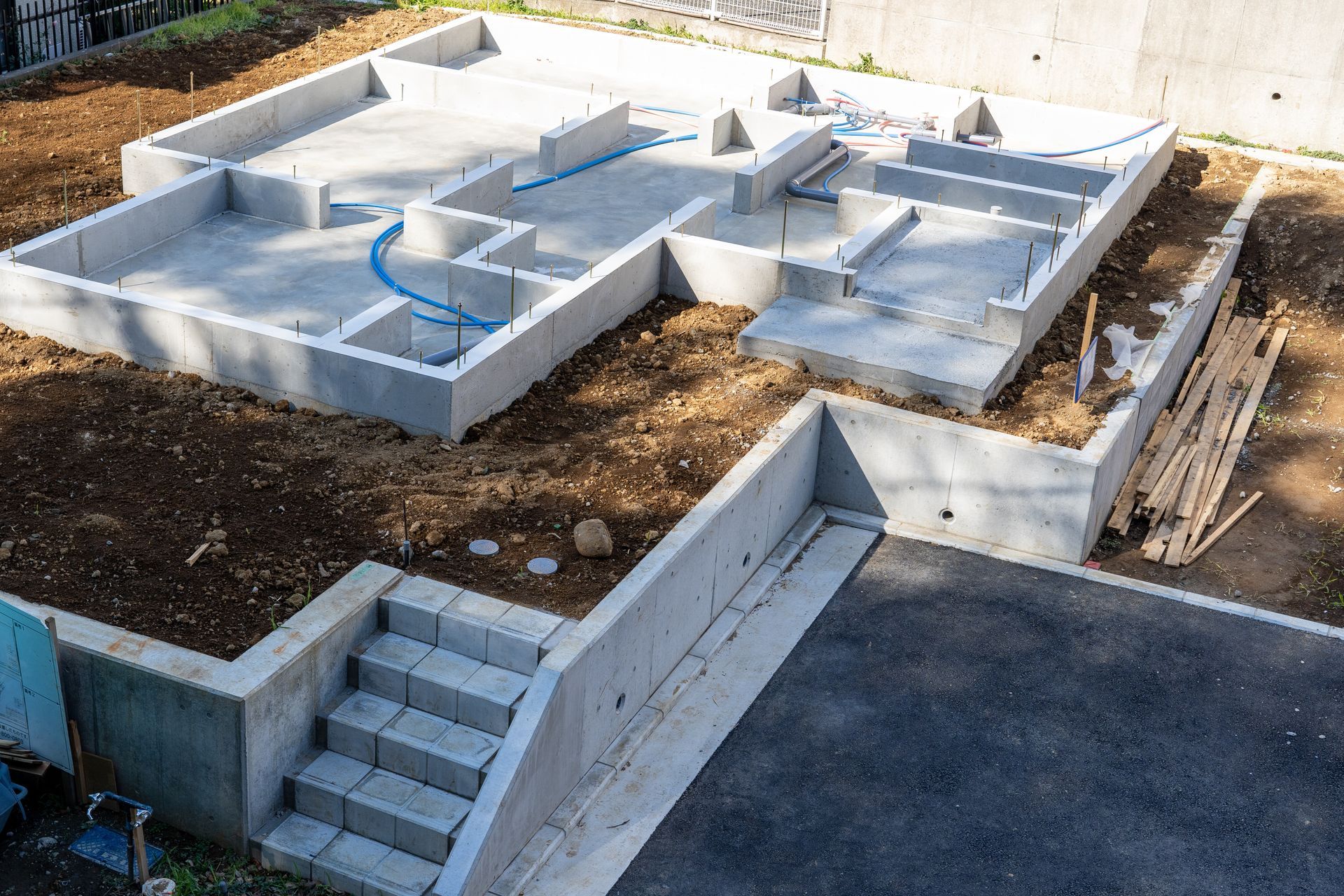 Fondations en béton en construction avec escaliers, murs et canalisations bleues sur un terrain en terre battue
