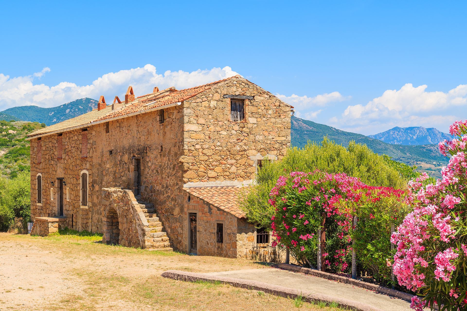 Maison rustique en pierre avec des buissons en fleurs, des montagnes et un ciel bleu dans un cadre champêtre ensoleillé