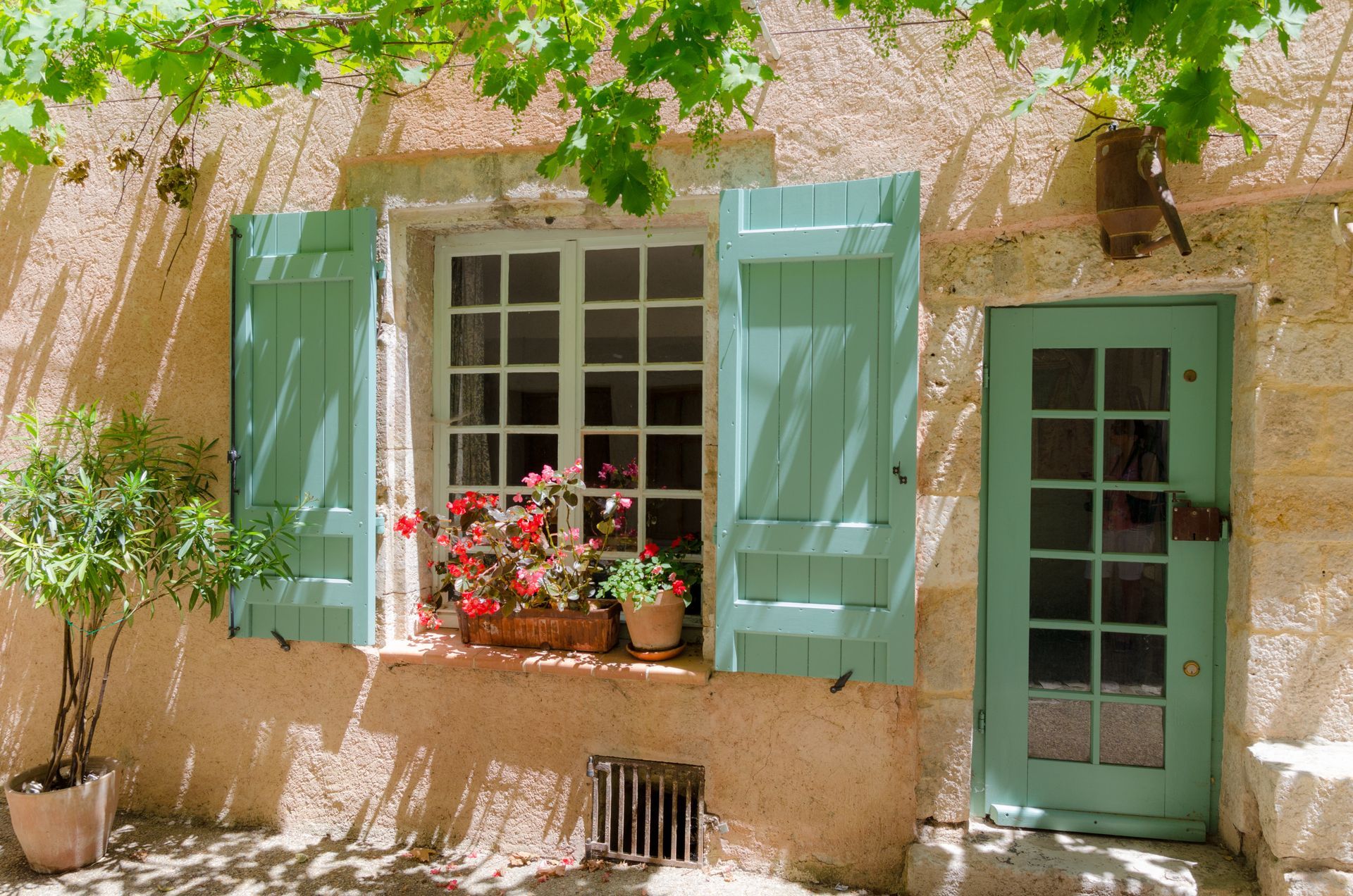 Mur en stuc beige baigné de soleil, avec des volets verts, des jardinières de fleurs rouges et une porte verte vitrée.