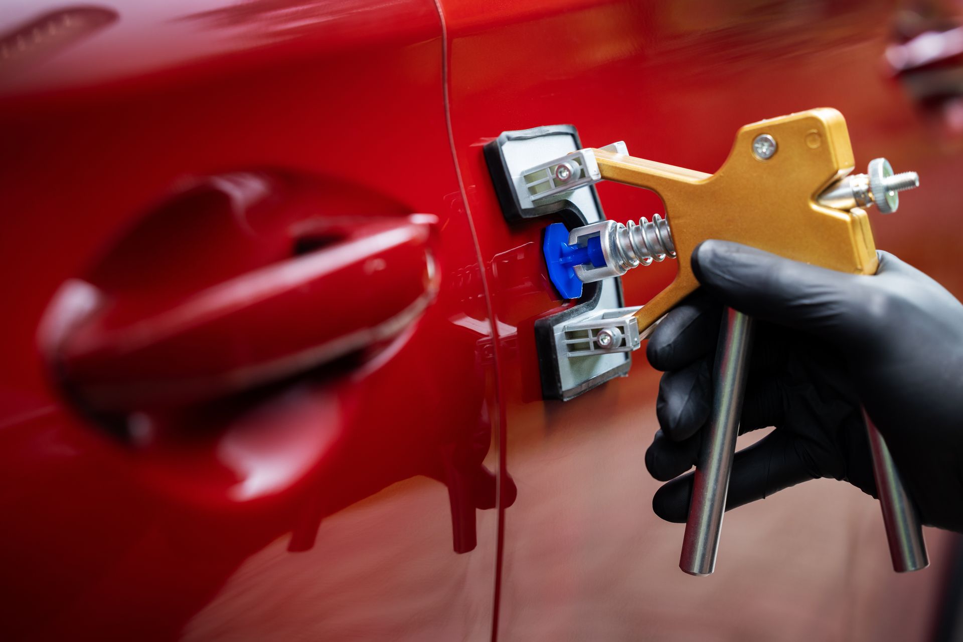 Une personne portant un gant utilise un outil de débosselage sur une portière de voiture rouge.
