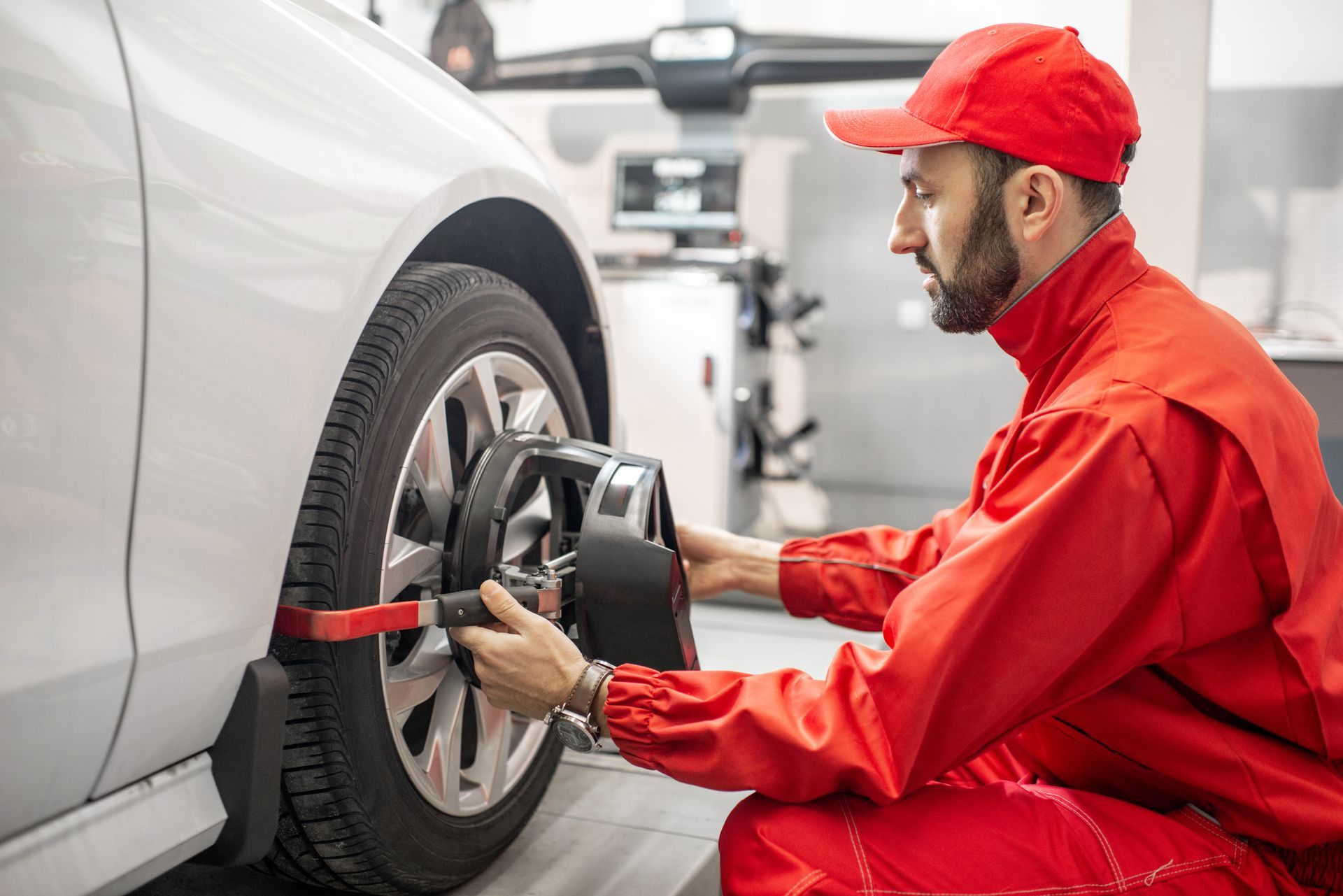 Un mécanicien en combinaison rouge règle l'équipement de géométrie des roues d'une voiture blanche dans un garage.