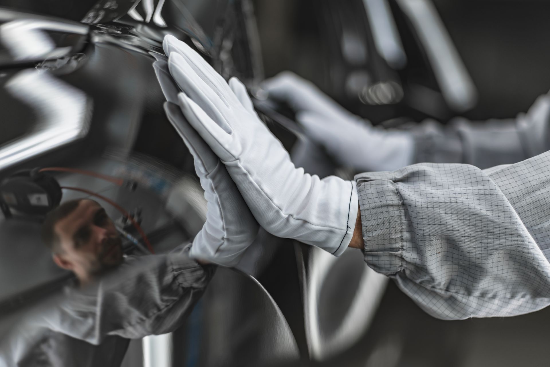 Une main gantée de blanc touche une surface métallique brillante, reflétant une personne.