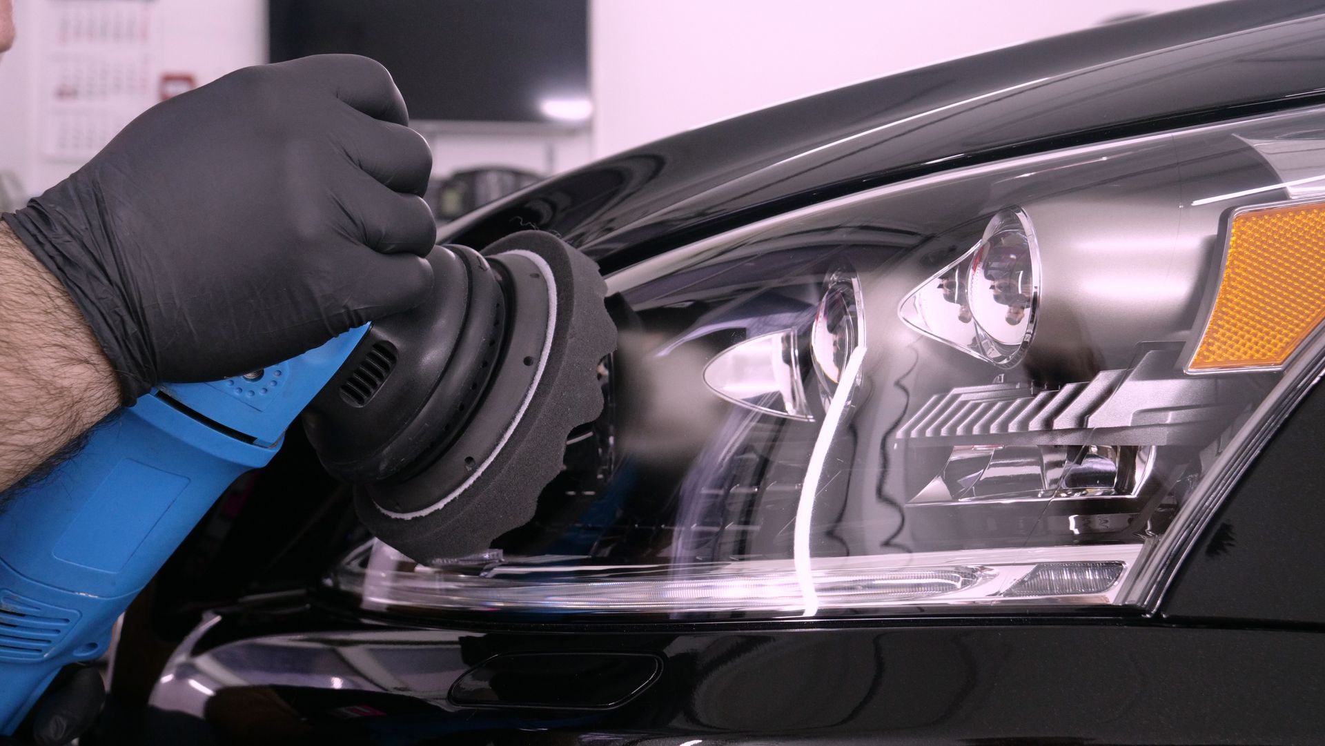 Une personne portant des gants noirs utilise une polisseuse bleue pour polir le phare d'une voiture.