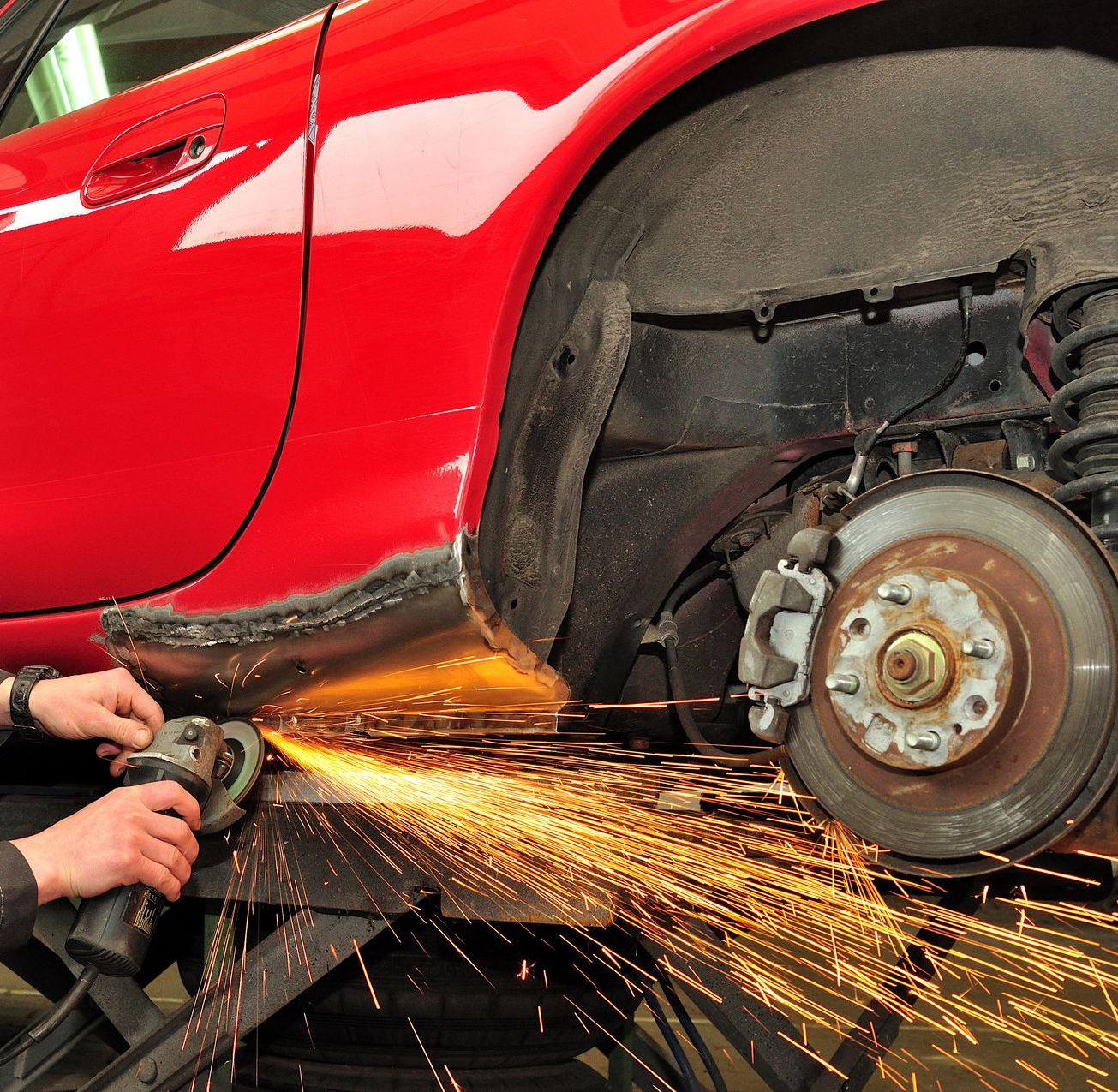 Une personne utilise une meuleuse, créant des étincelles, sur le côté d'une voiture rouge, près de la roue.