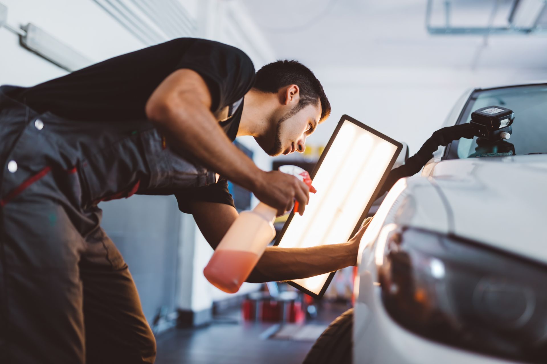 Un carrossier examine une bosse sur une voiture blanche à l'aide d'une lampe torche et d'un vaporisateur dans un garage.