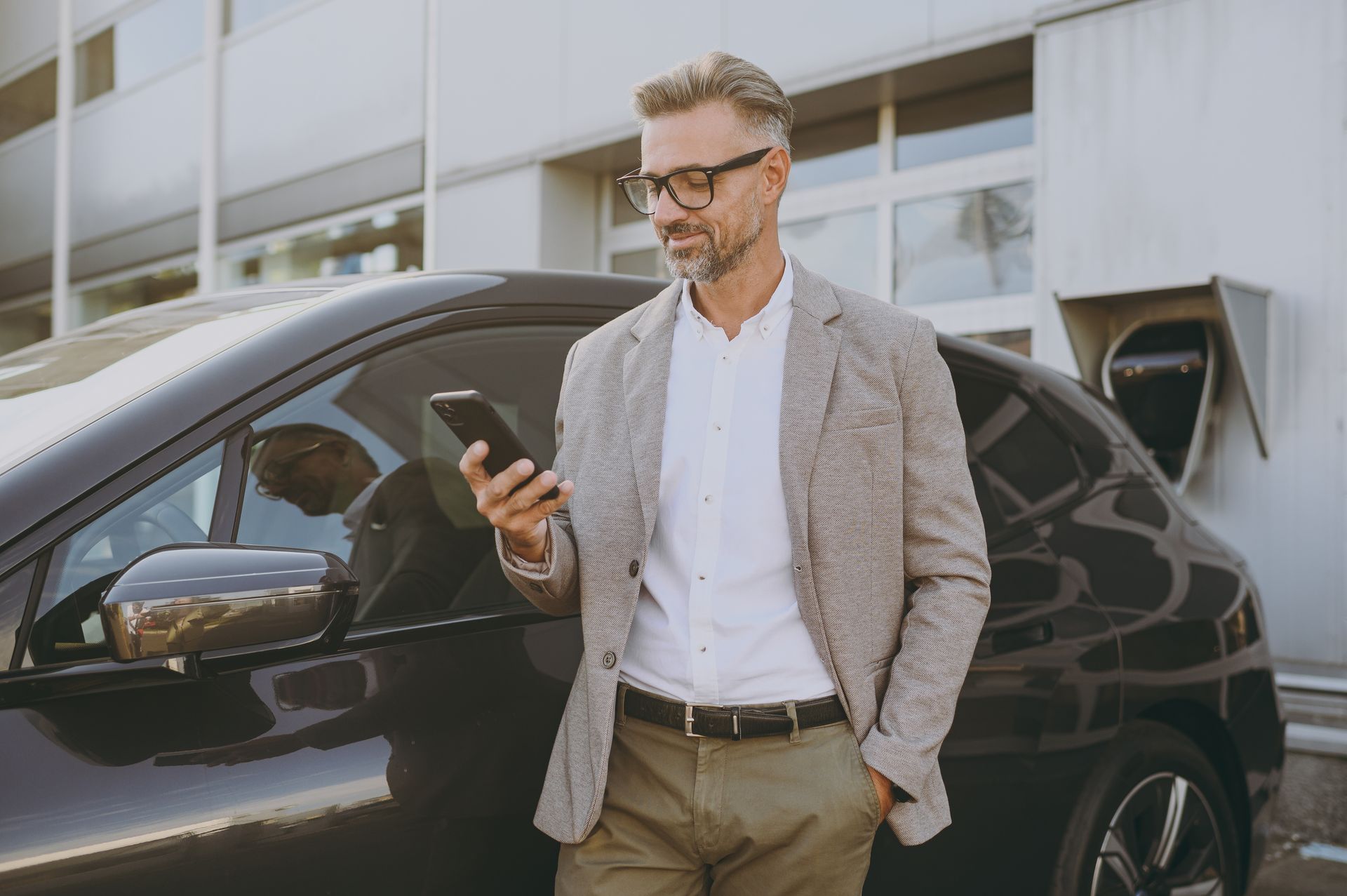 Un homme en blazer utilise son téléphone, appuyé contre une voiture électrique noire près d'une borne de recharge.