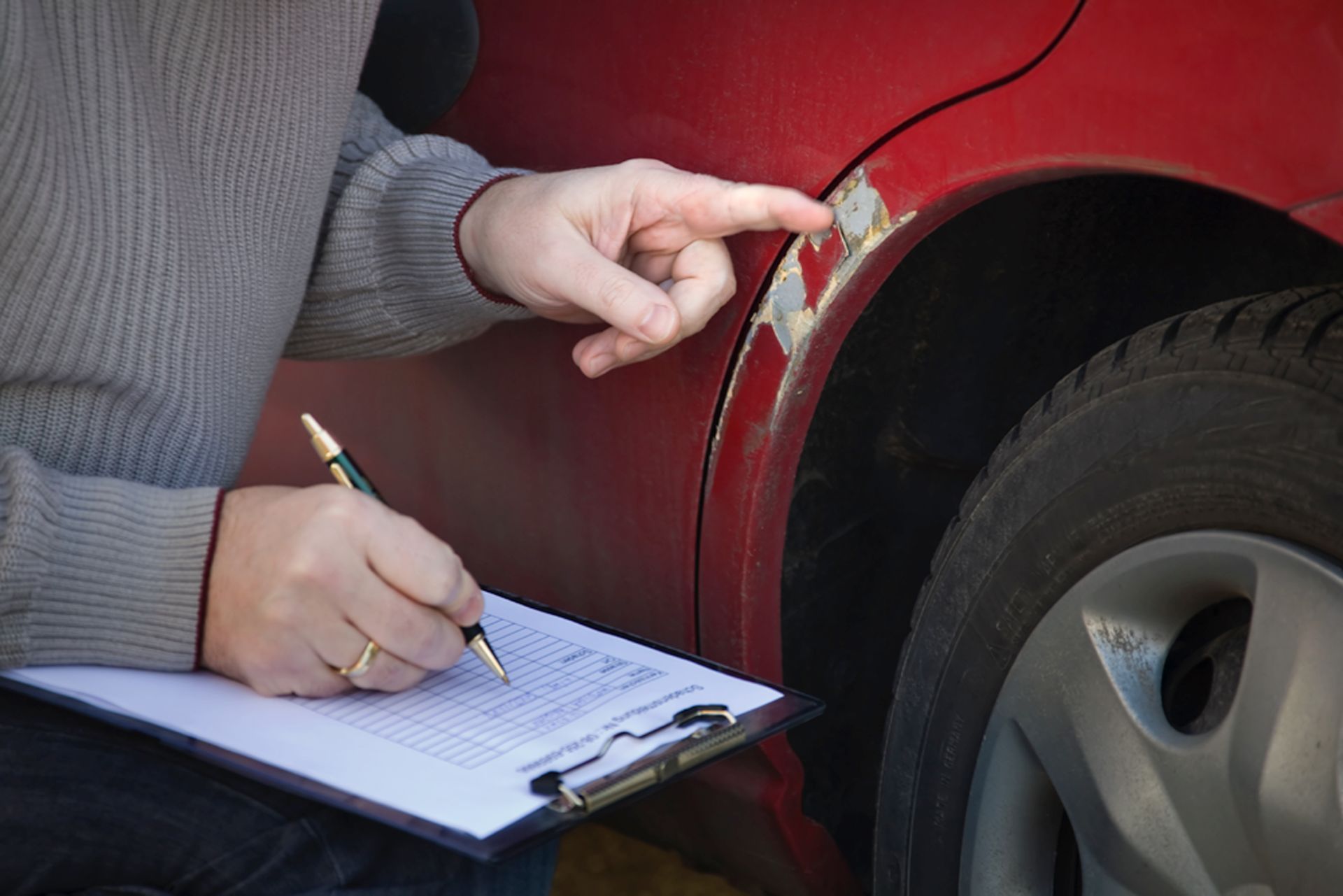 Une personne inspecte les dommages causés à une voiture, montrant du doigt les rayures sur la peinture, prenant des notes sur un bloc-notes.