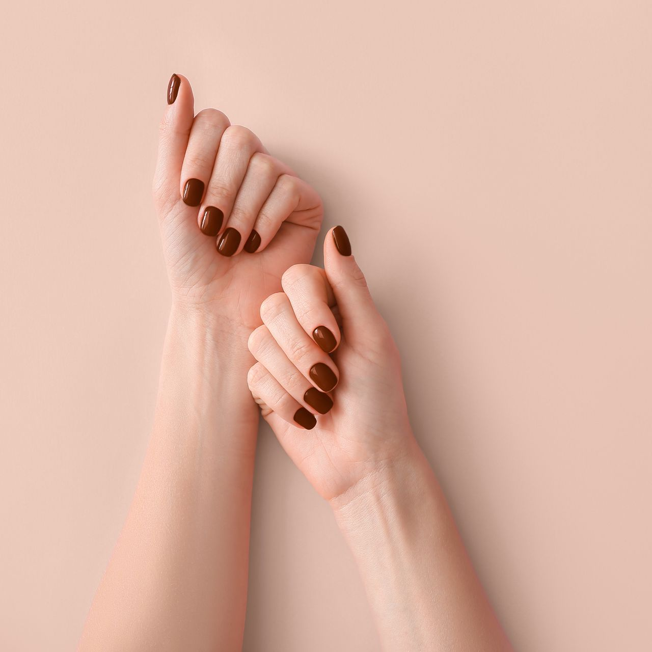 Mains avec vernis à ongles marron foncé sur fond pêche.