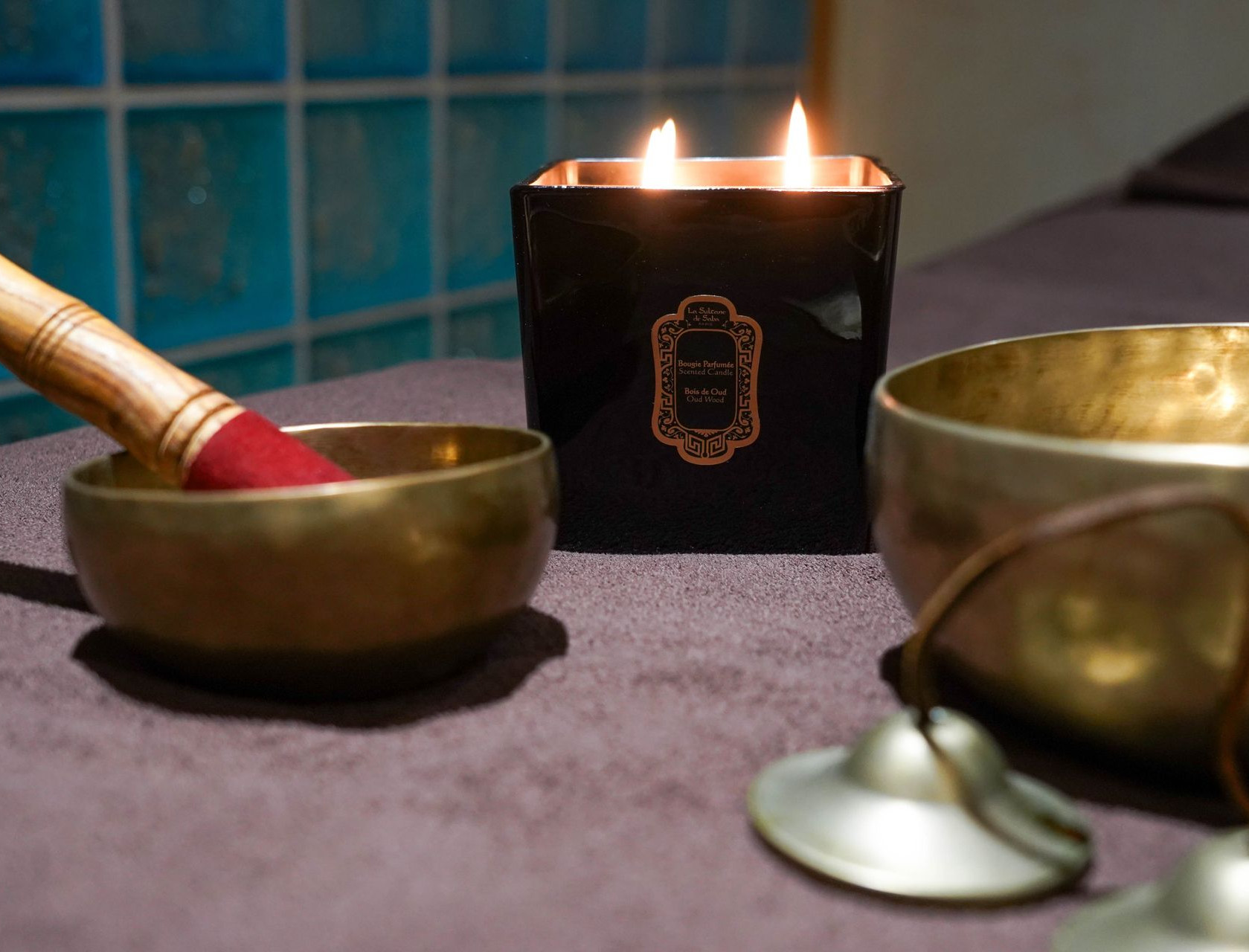 Bols chantants dorés, bougie et maillet sur une table de spa avec fond de carrelage bleu.