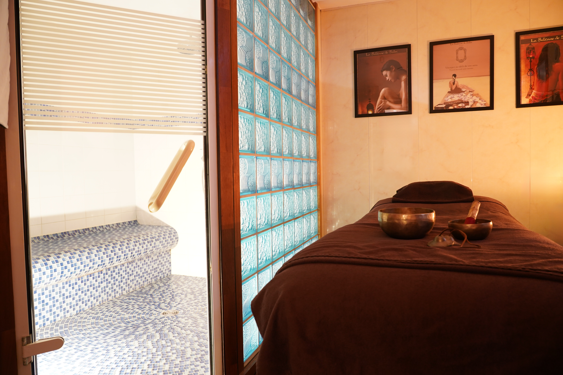 Salle de massage avec mur en blocs de verre, table de massage et petit espace carrelé.
