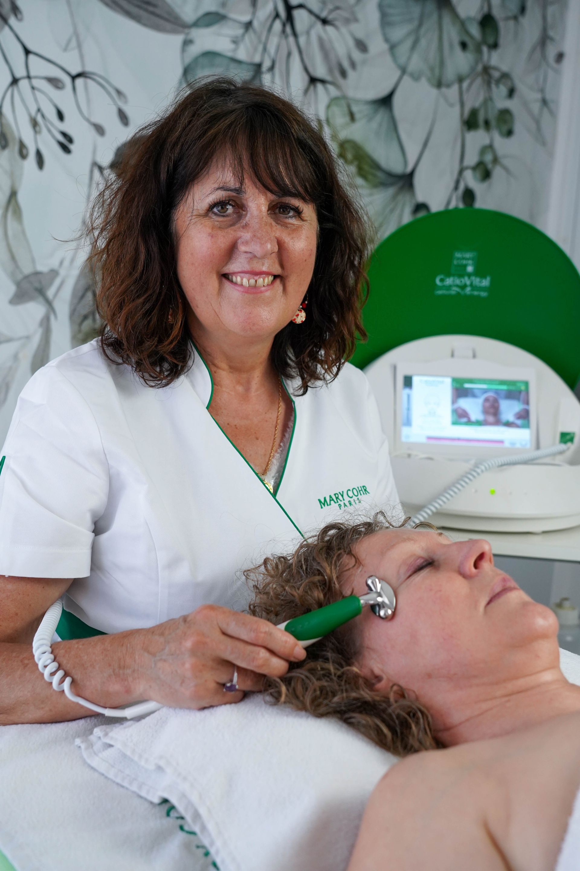 Une femme en uniforme blanc utilise un appareil facial sur une autre femme dans une salle de traitement.