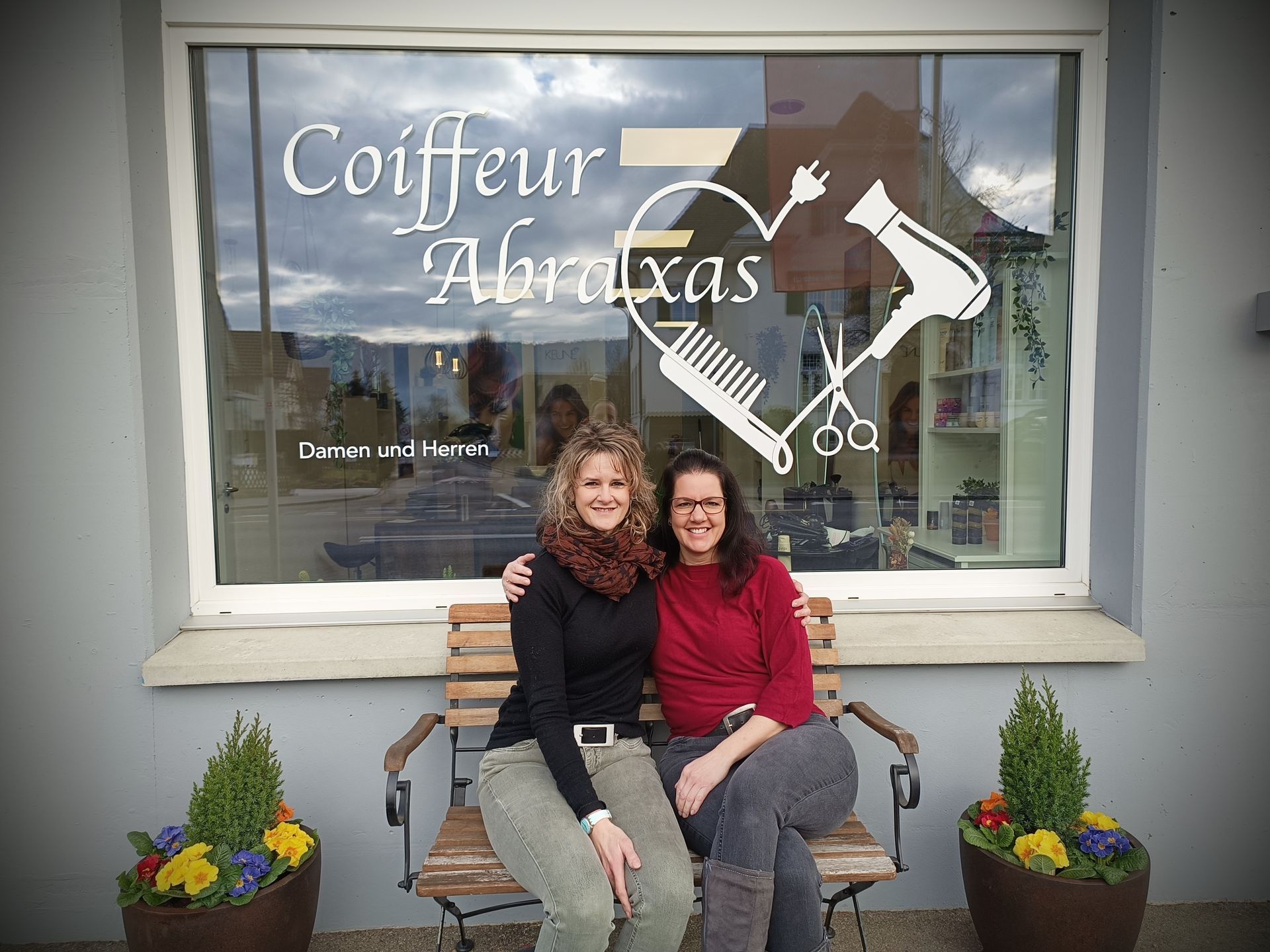Zwei Frauen sitzen auf einer Bank vor einem Fenster mit der Aufschrift „Coiffeur Abraxas“