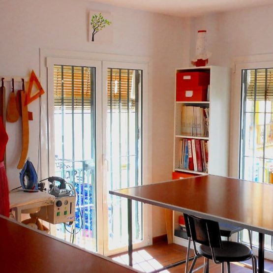 Comedor luminoso con mesa de madera, sillas negras, estanterías y puertas de cristal que dan a un balcón.