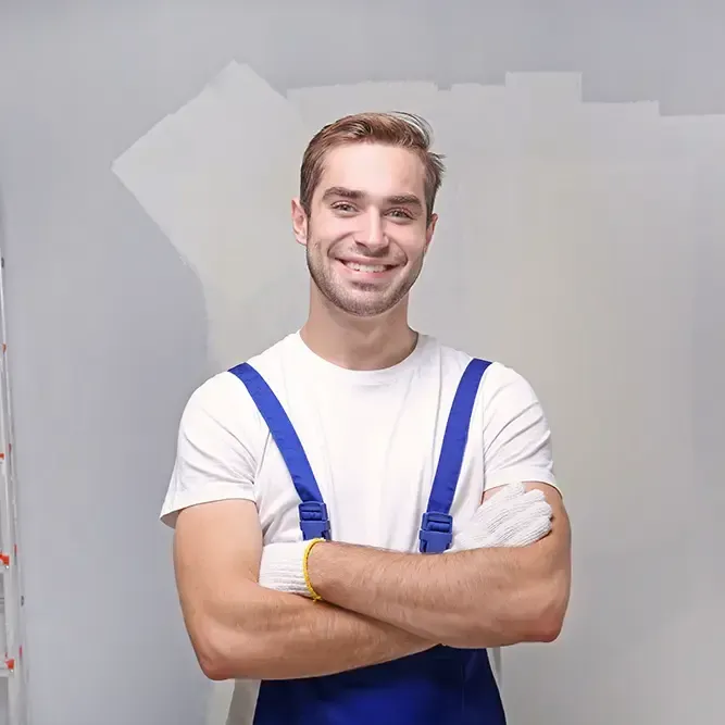 Ein Mann in blauem Overall und weißen Handschuhen lächelt mit verschränkten Armen vor einer bemalten Wand.