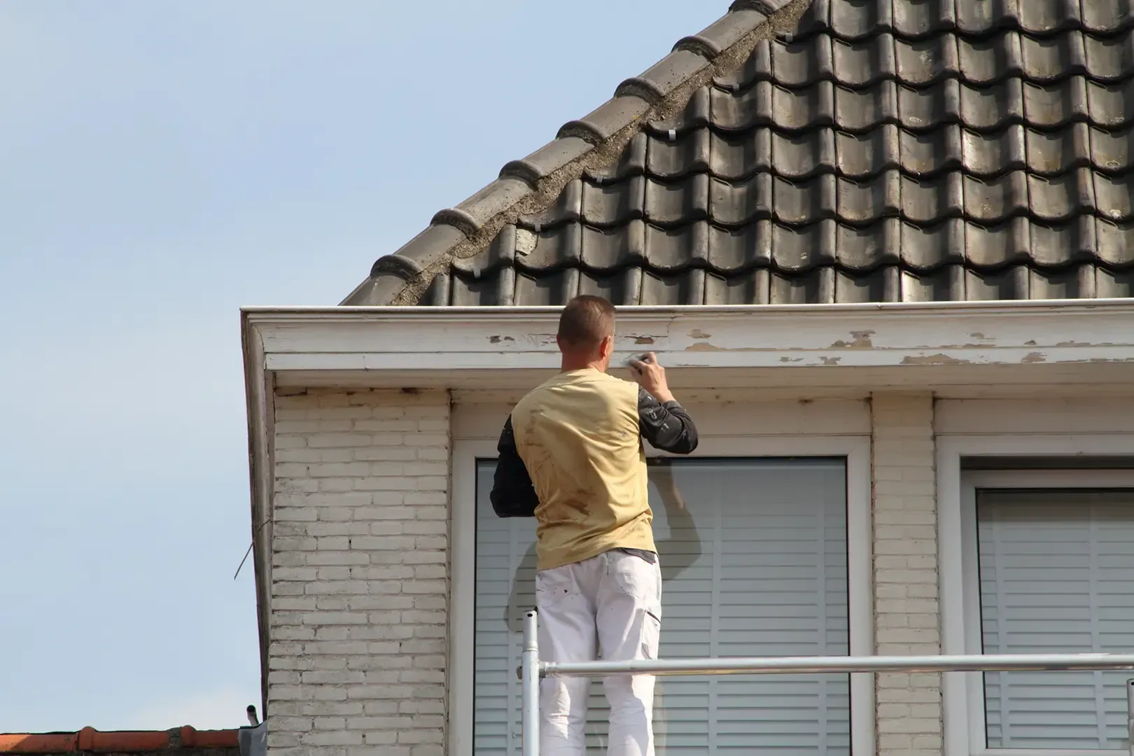 Eine Person, die auf einem Gerüst steht, streicht die Zierleisten eines Hauses mit Ziegeldach.