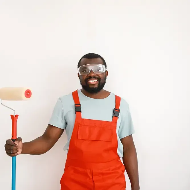 Eine Person mit Farbrolle, Schutzbrille und orangefarbenem Overall steht vor einer weißen Wand.